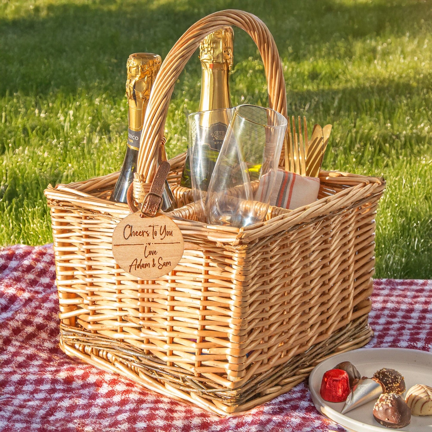 A personalised wicker wine carrier basket is in a picnic outdoor spot. Featuring a personalised cherry wood veneer tag
