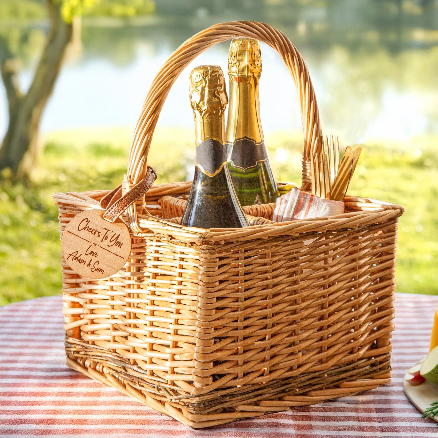 A personalised wicker wine carrier basket is in a picnic outdoor spot. Featuring a personalised cherry wood veneer tag