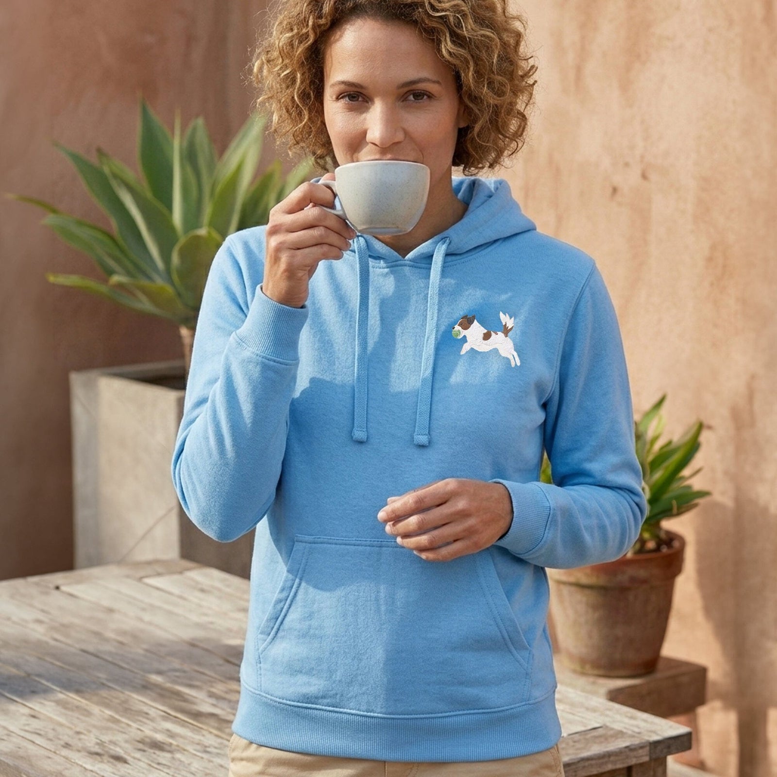 A woman with curly hair wearing a light blue hoodie with a small white and brown dog graphic on the left chest, holding a white mug in front of her face. She is standing outdoors next to a wooden table with potted plants.