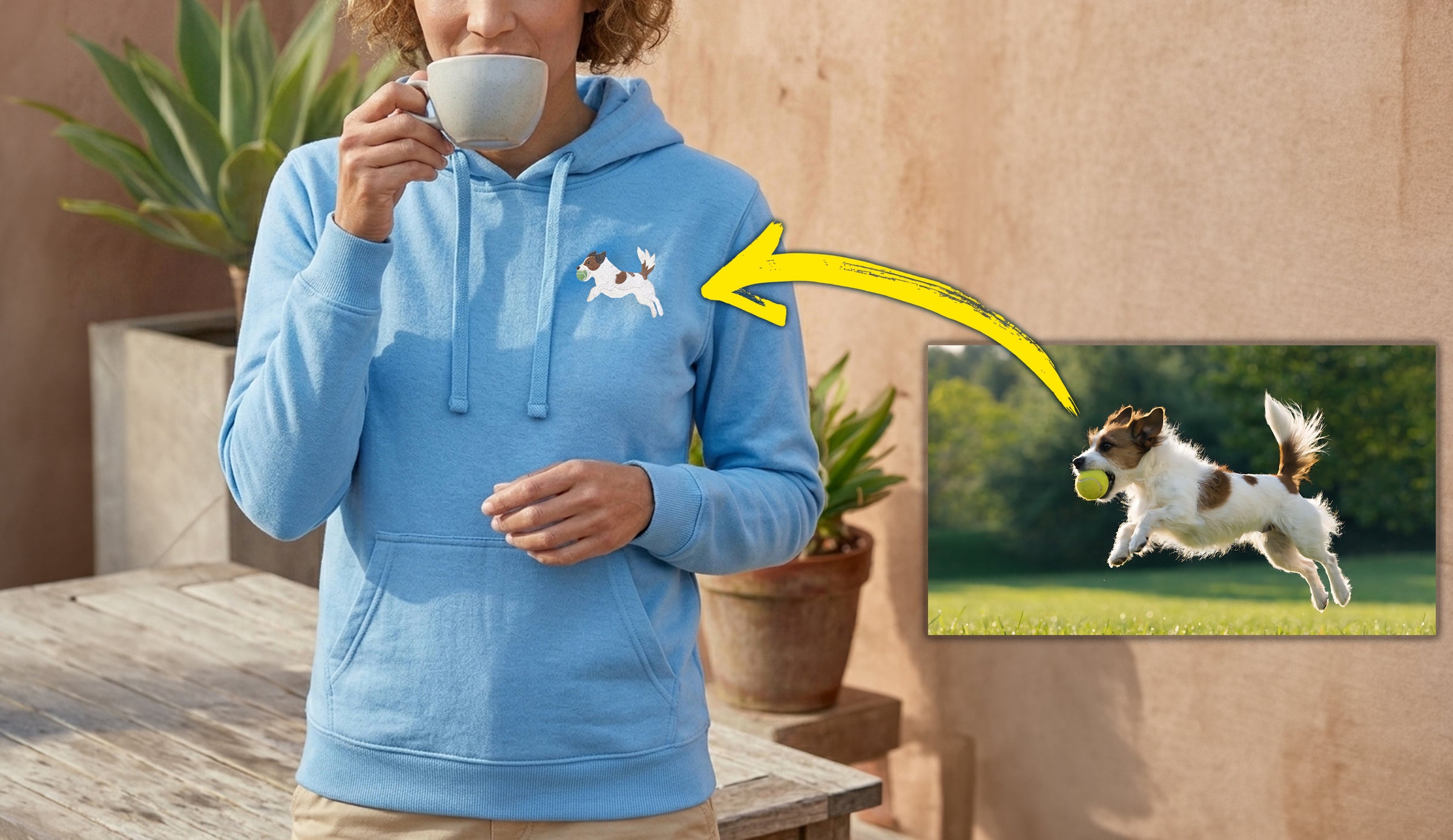 A woman wearing a light blue hoodie with a small white and black dog graphic on the left chest, holding a white mug. A yellow arrow points from the hoodie graphic to an inset photo of a jumping dog holding a tennis ball.
