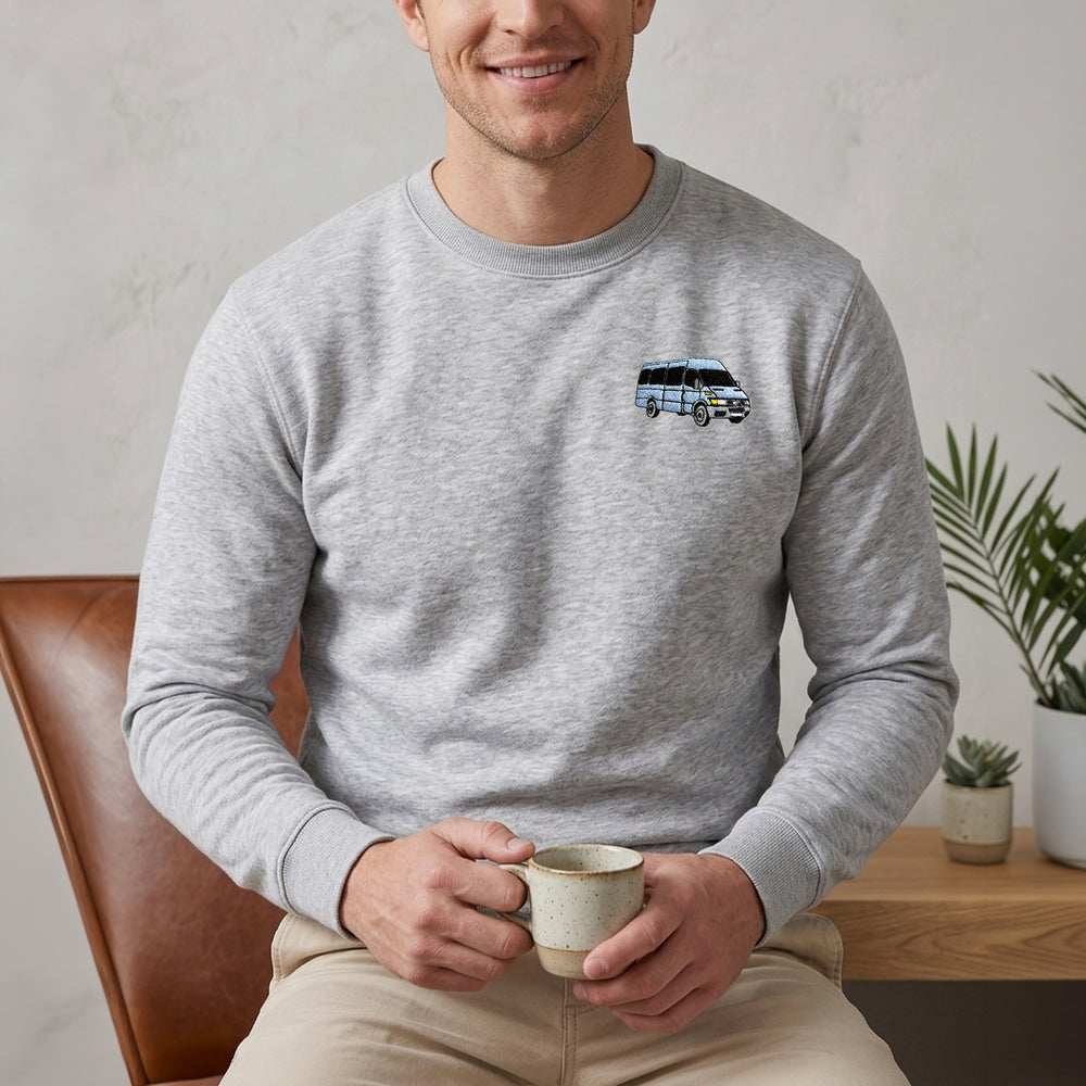 A man wearing a light gray crewneck sweatshirt with a small graphic of a blue and white van on the left chest, sitting in a brown leather chair and holding a ceramic mug.