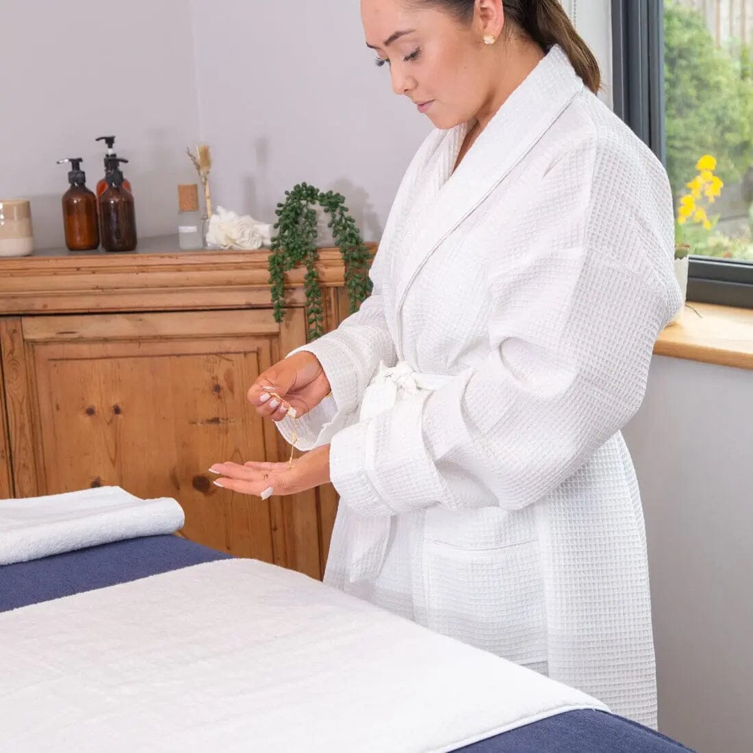 young women removing jewellery, wearing a spa robe and standing next to a couch with a cover on it  