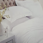 White bedding with two pillows on a bed featuring a tufted beige headboard, with a white bedside table and vase of flowers in the foreground.