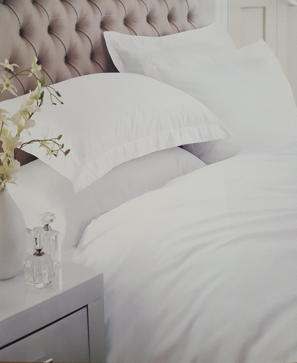 White bedding with two pillows on a bed featuring a tufted beige headboard, with a white bedside table and vase of flowers in the foreground.