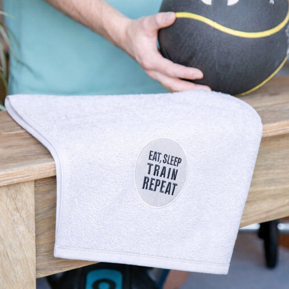 White towel with 'EAT.SLEEP TRAIN REPEAT' text on a wooden surface, held by a person with a medicine ball.