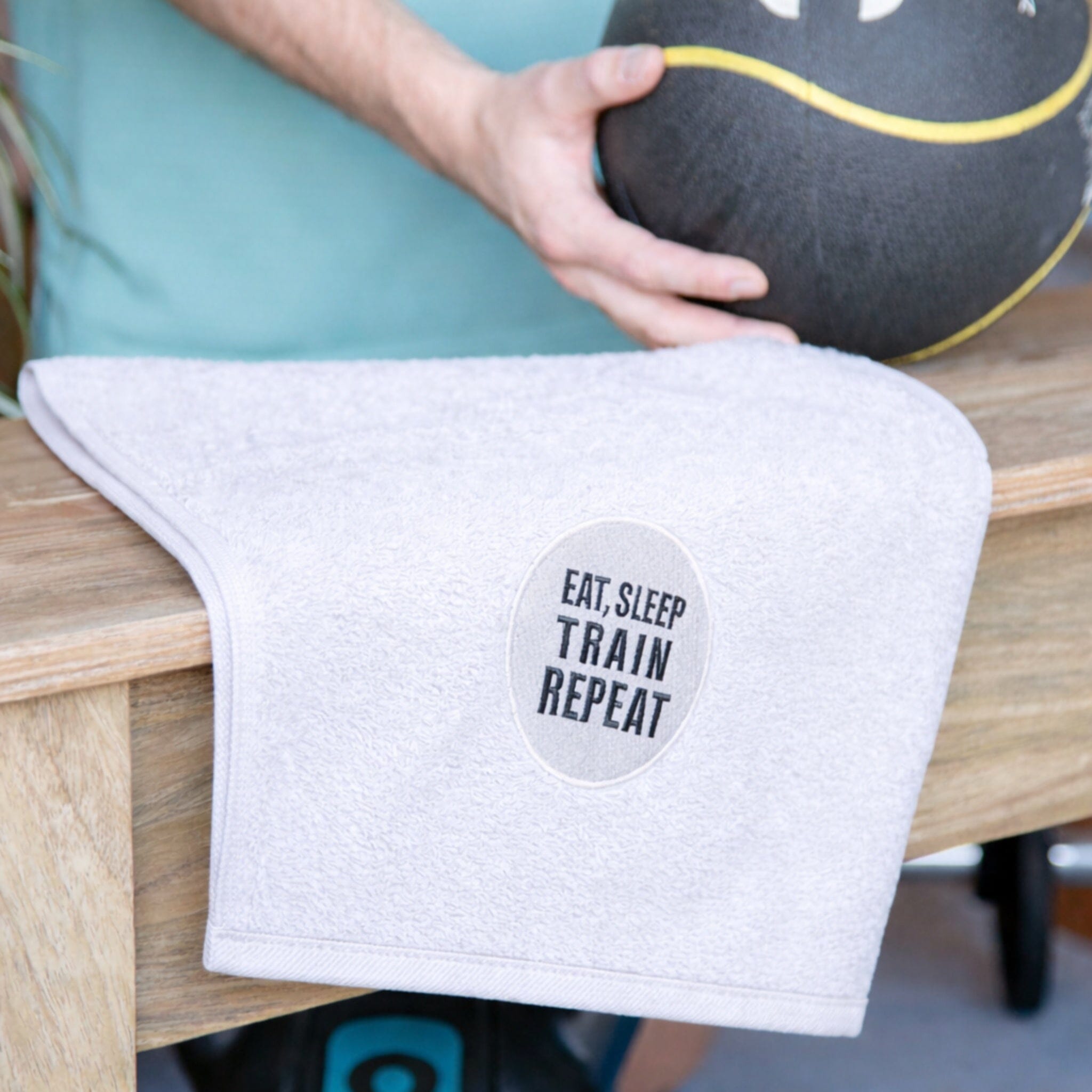White towel with 'EAT.SLEEP TRAIN REPEAT' text on a wooden surface, held by a person with a medicine ball.