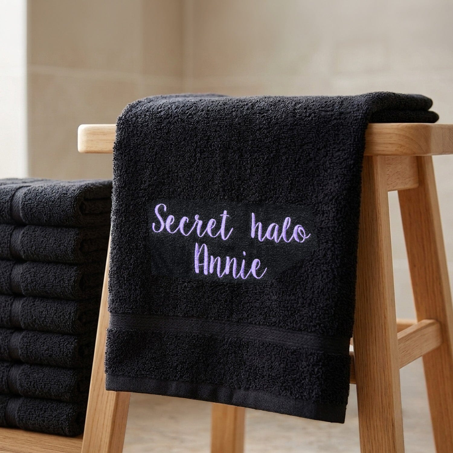 A Personalised black hair dressing towel embroidered in lilac thread, draped over a wooden stool next to a stack of folded towels.