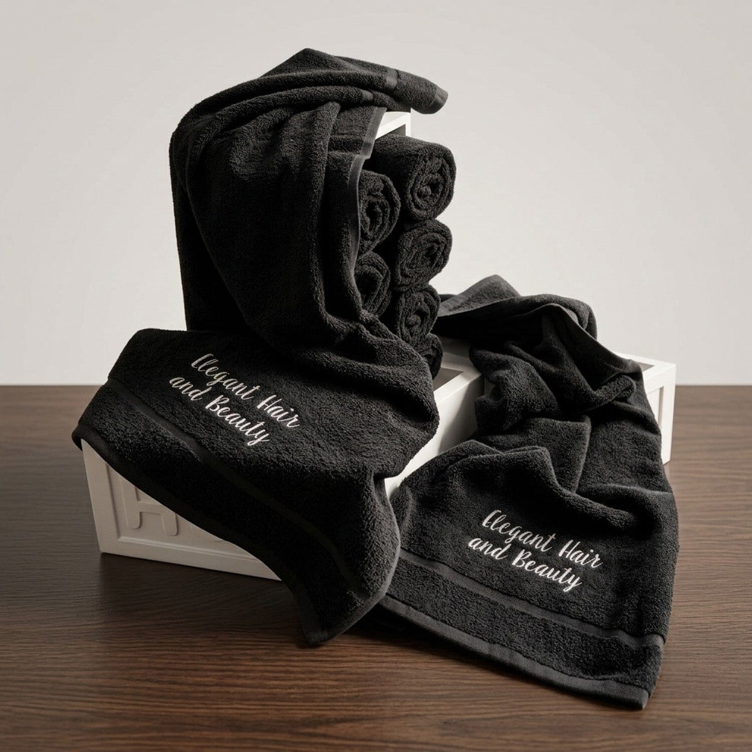 A set of 10 black cotton Hair dressing towels, some rolled, a couple embroidered with 'Elegant Hair and Beauty' in white thread.