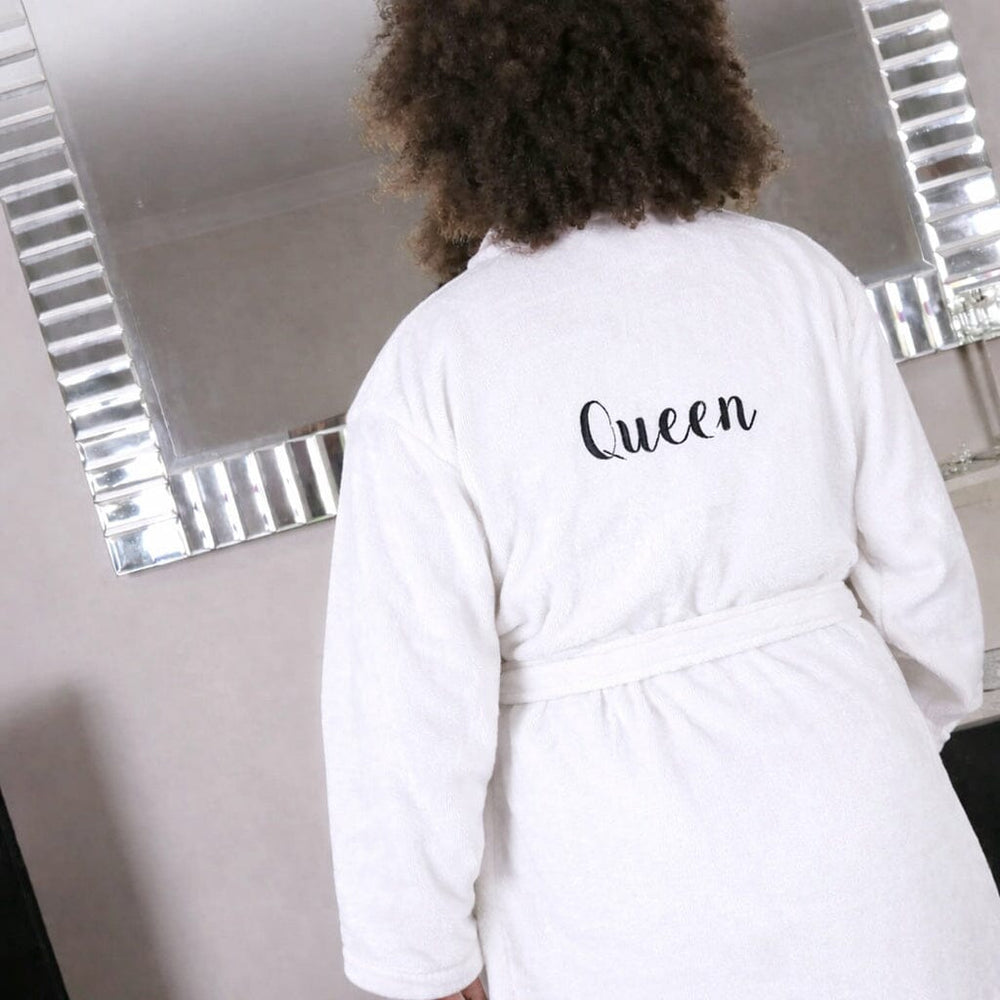 Person wearing a white robe with 'Queen' printed on the back, standing in front of a mirror.
