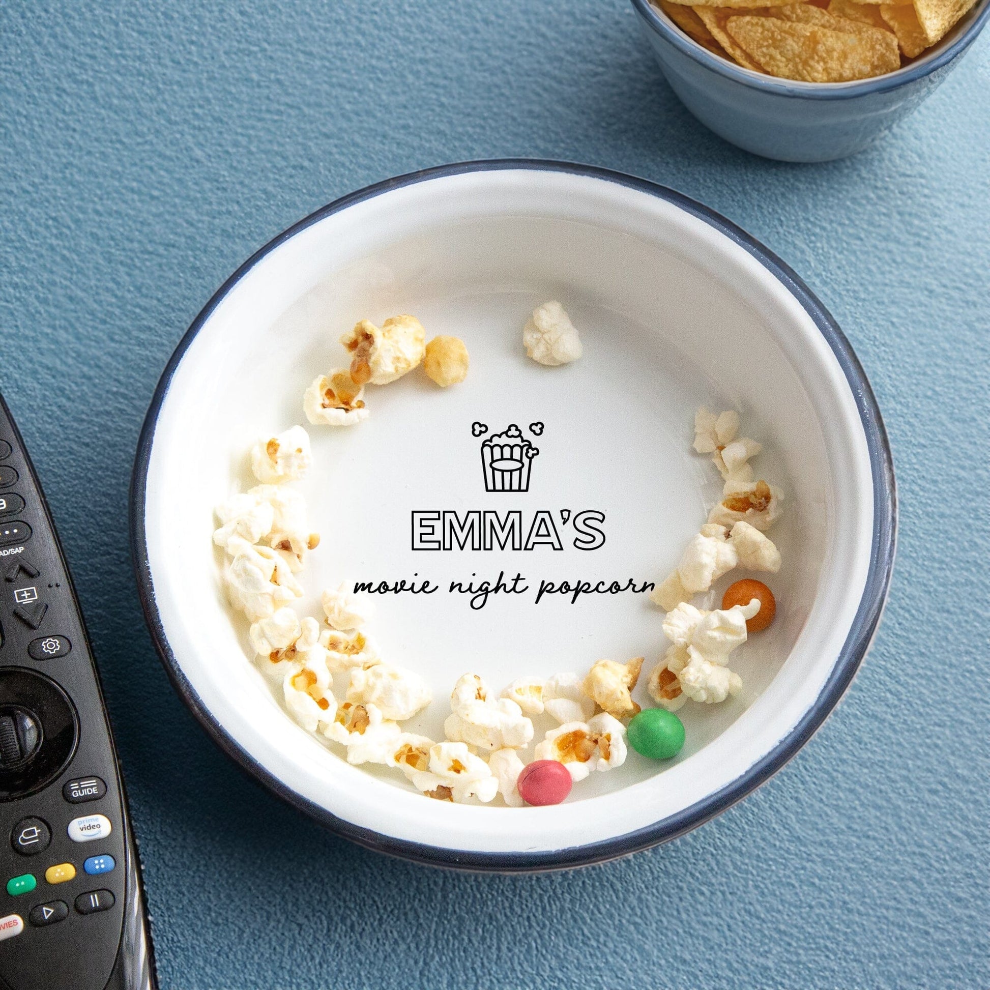 Personalised Children's Popcorn Enamel Bowl on the table. A remote popcorns and crisps around