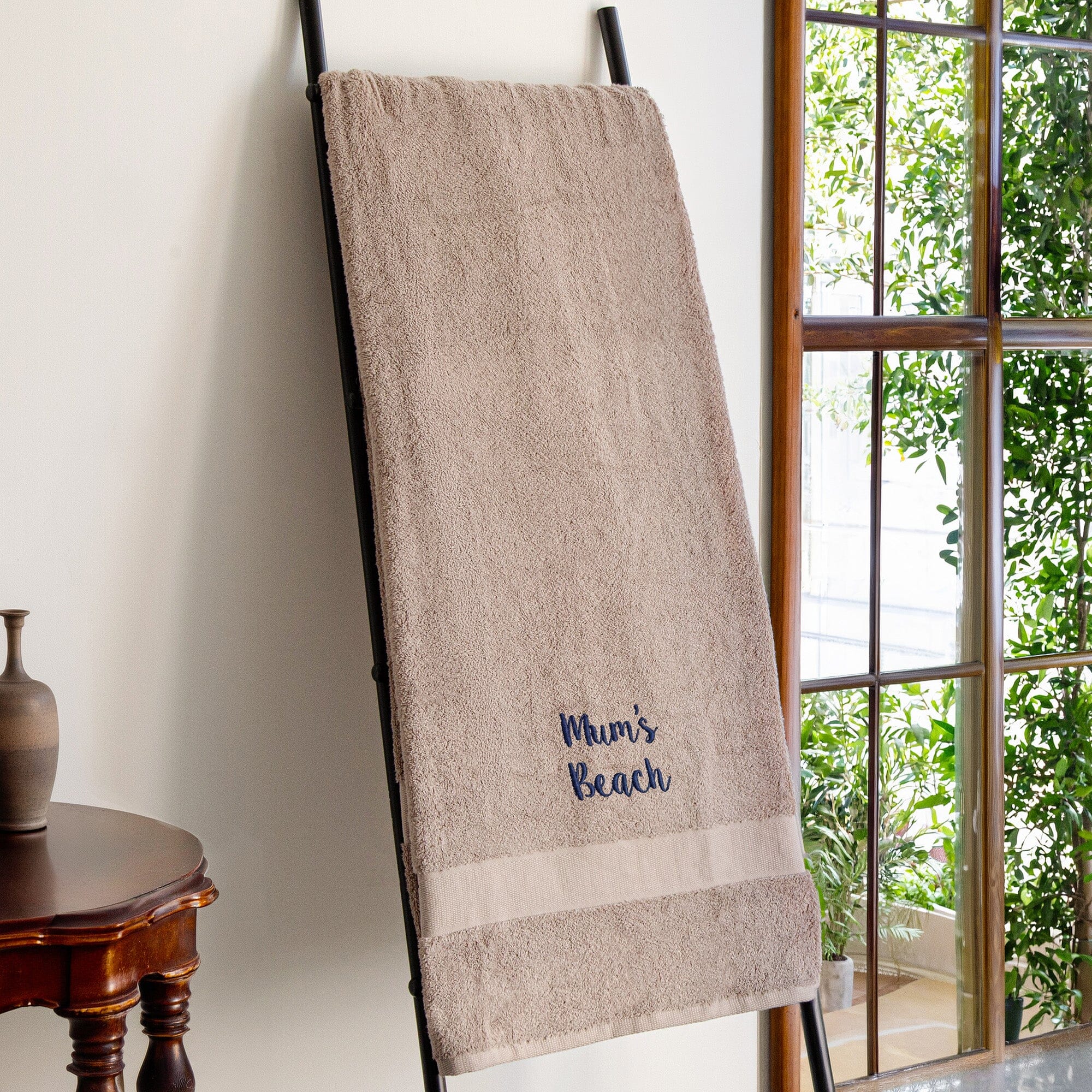 A beige bath towel with a navy blue embroidered 'Mum's Beach' design, hanging on a black metal stand in a bright room next to a window.