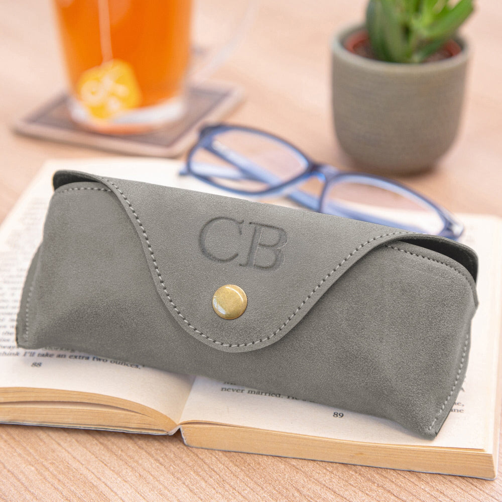 A grey faux-leather personalised eyeglasses case with a snap closure and debossed 'CB' monogram, resting on an open book with blue-framed glasses and a blurred drink in the background.