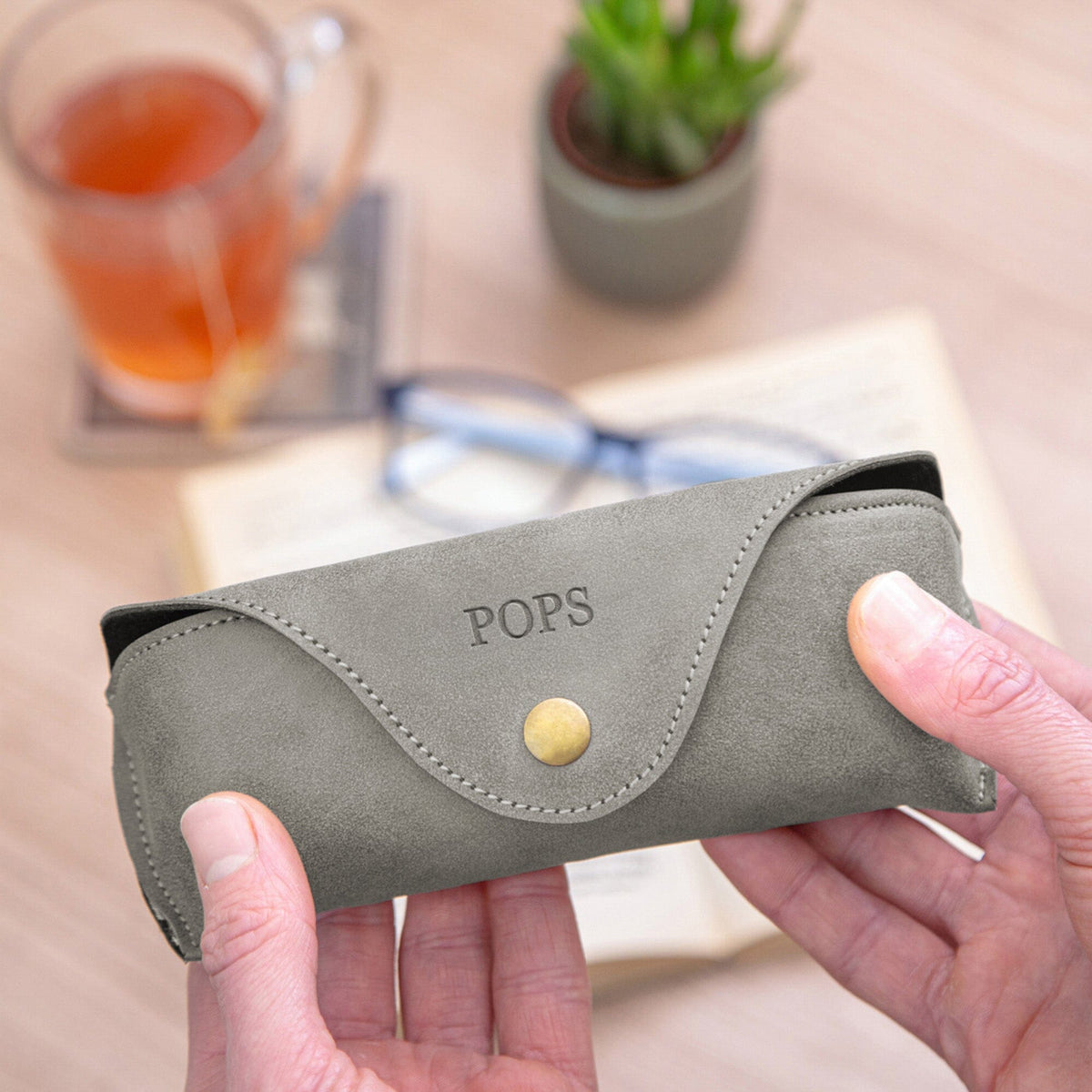 A person holds a personalised slate faux-leather glasses case with a snap button closure and the brand name 'POPS' debossed on the front. The background shows a wooden desk with a glass of tea, a small potted plant, and a pair of eyeglasses.
