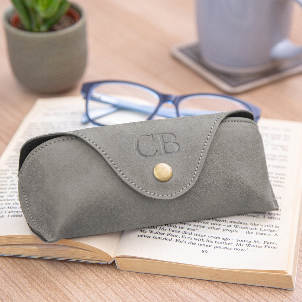 A grey faux leather personalised eyeglasses case with a debossed 'CB' monogram and gold snap closure, resting on an open book with blue eyeglasses and a small potted plant in the background.