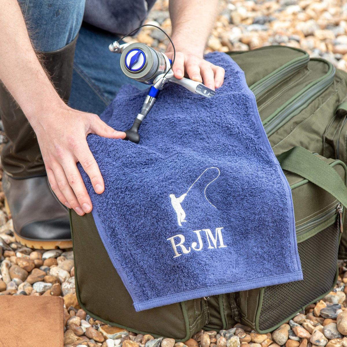 A navy-coloured personalised fishing towel laid on a fishing bag on beach pebbles, featuring the initials 'RJM' and fishing silhouette embroidered. 