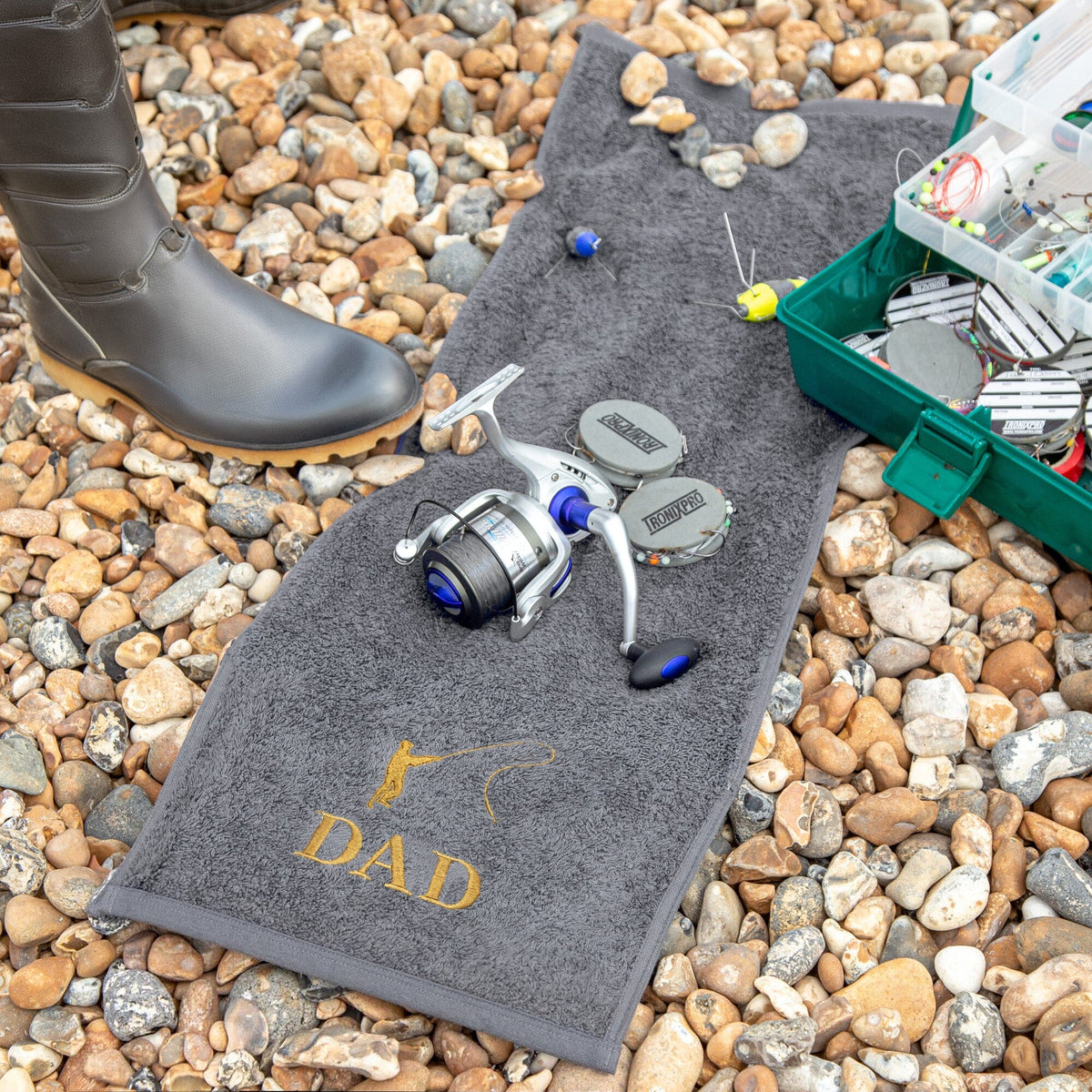 A slate-coloured personalised fishing towel laid on a bed of pebbles, featuring the name 'DAD' in gold embroidery. A black boot, a silver and blue spinning reel, fishing hooks, and a green tackle box are arranged around the towel.
