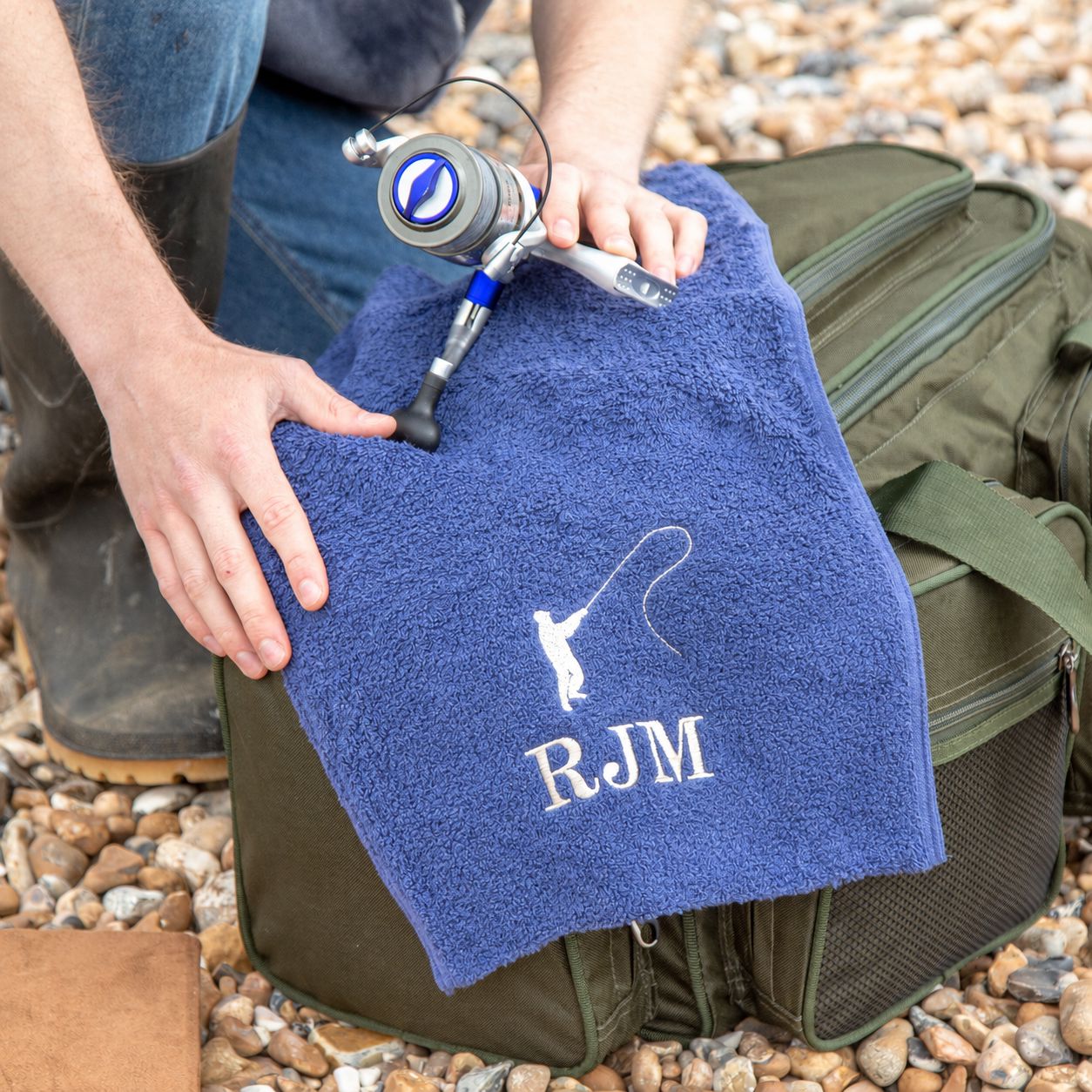 A person outdoors on a pebble beach holds a blue terry towel with a white fishing rod and line graphic and the embroidered initials 'RJM' over an olive green tackle bag.