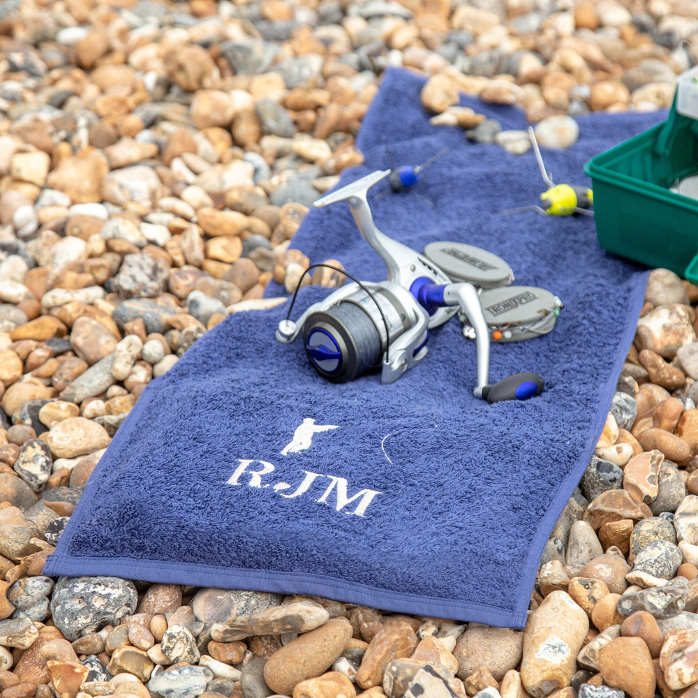 A navy-coloured personalised fishing towel laid on a bed of pebbles, featuring the initials 'RJM' and fishing silhouette embroidered. Fishing accessories arranged around the towel.