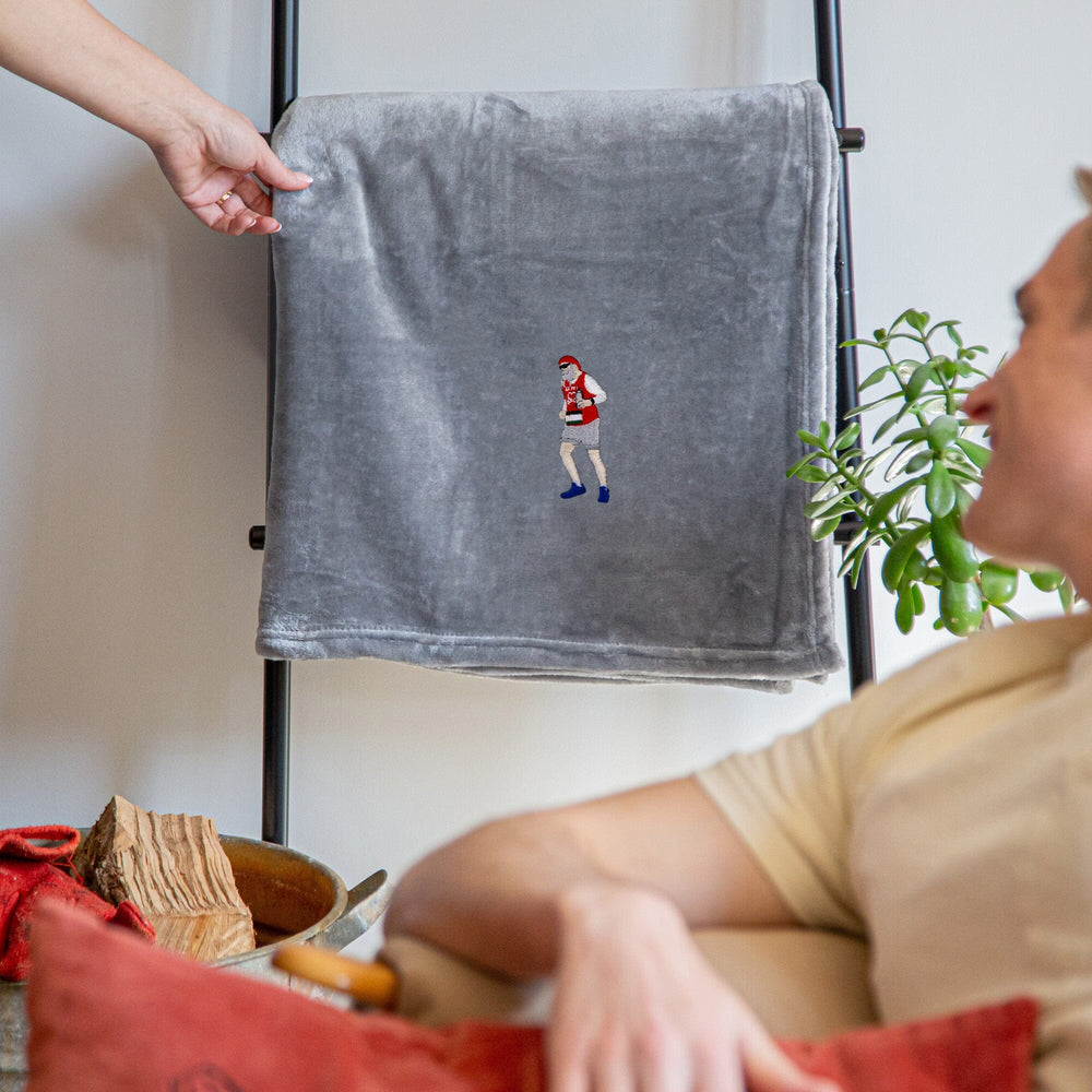 A grey fleece throw blanket with a small embroidered boxer character is draped over a black metal stand in a cozy indoor setting, with a person holding the top corner and another person sitting nearby.