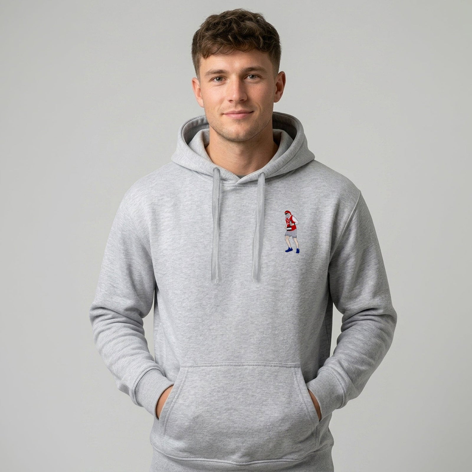 A man wearing a light gray heather pullover hoodie with a small embroidered cartoon character on the left chest, standing against a solid gray studio background.