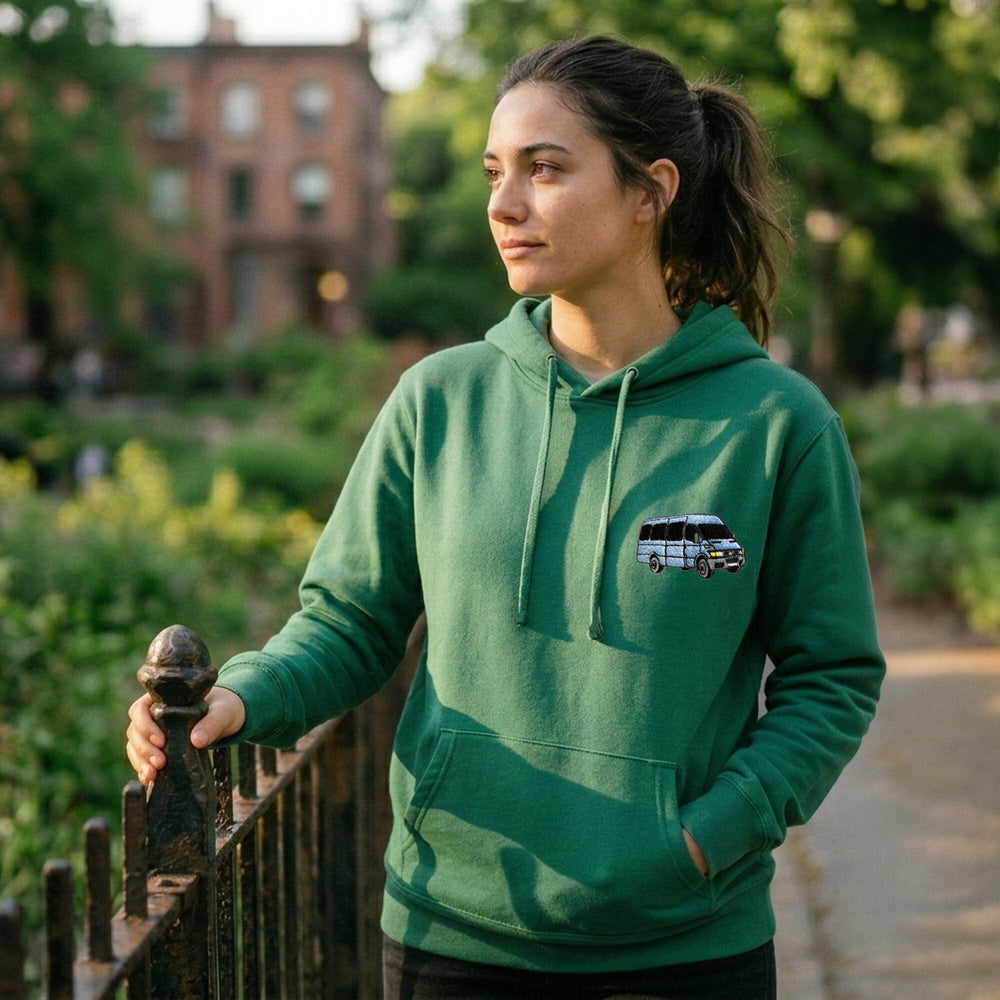 A woman wearing a green pullover hoodie with a small graphic of a white and blue van on the left chest, standing outdoors near a fence in a park-like setting.