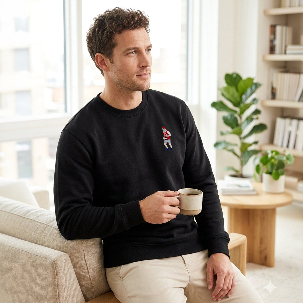 A man sitting on a beige couch in a bright, modern living room, wearing a black crewneck sweatshirt with a small embroidered hockey player on the left chest and holding a ceramic mug.