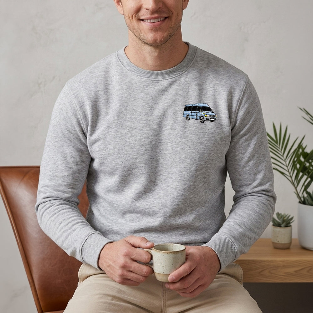 A man wearing a light gray crewneck sweatshirt with a small graphic of a blue and white van on the left chest, sitting in a brown leather chair and holding a ceramic mug.