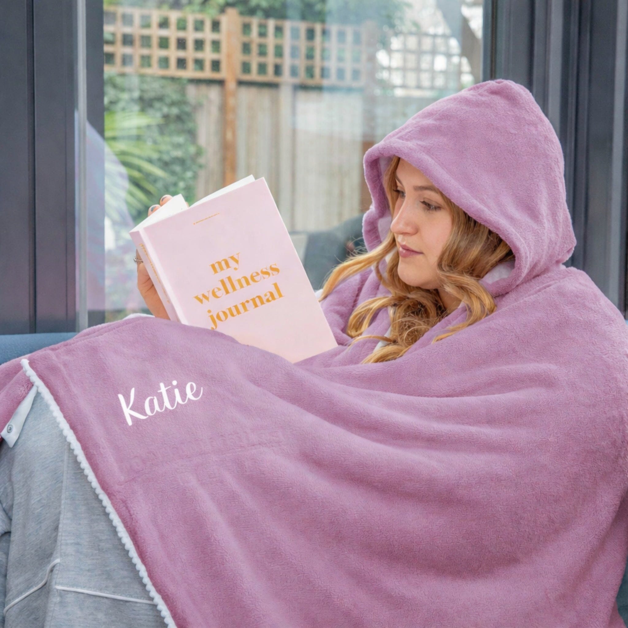 Person wrapped in a pink hooded blanket reading a 'my wellness journal' book.