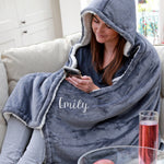 Person wrapped in a gray hooded blanket with 'Emily' embroidered on it, using a phone.