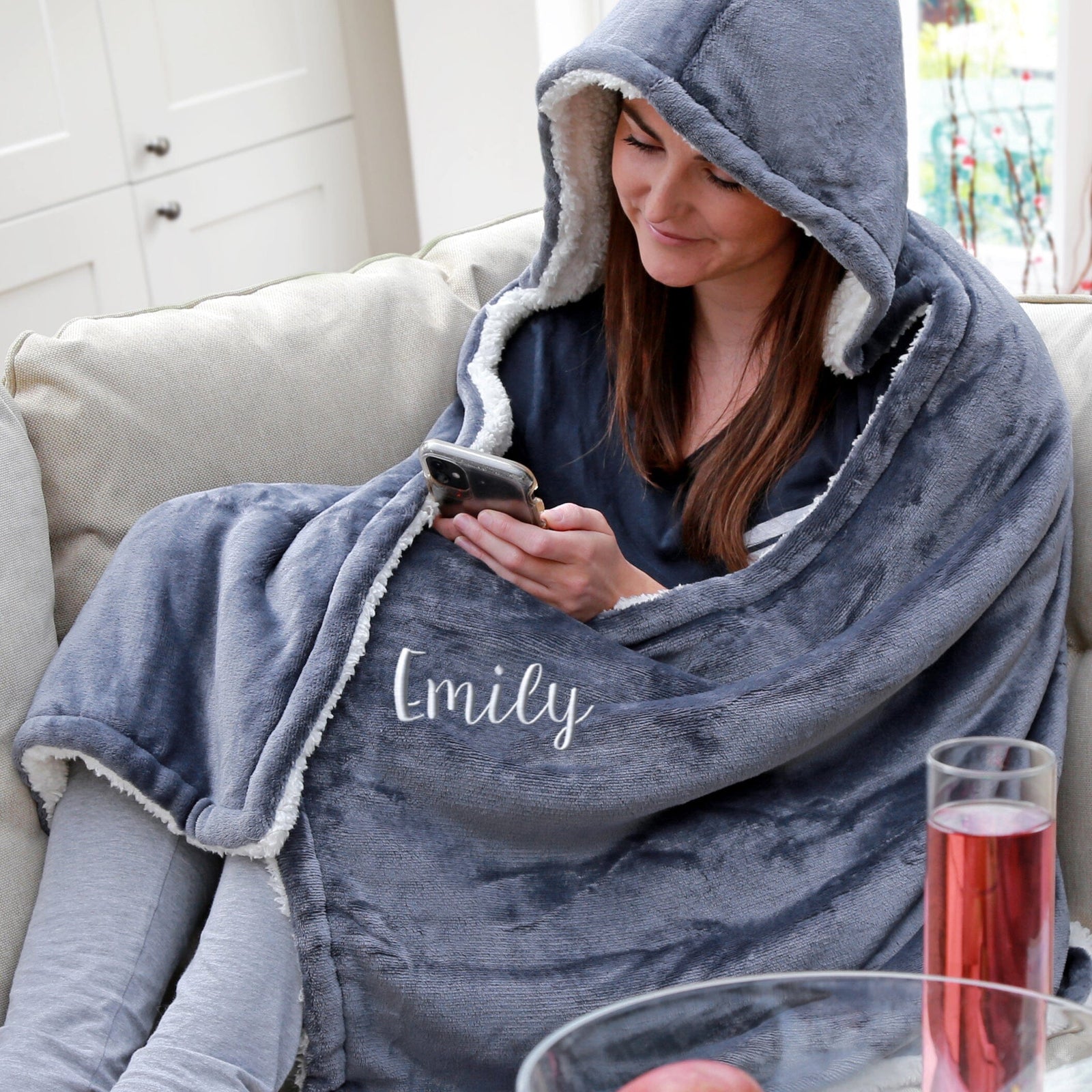 Person wrapped in a gray hooded blanket with 'Emily' embroidered on it, using a phone.