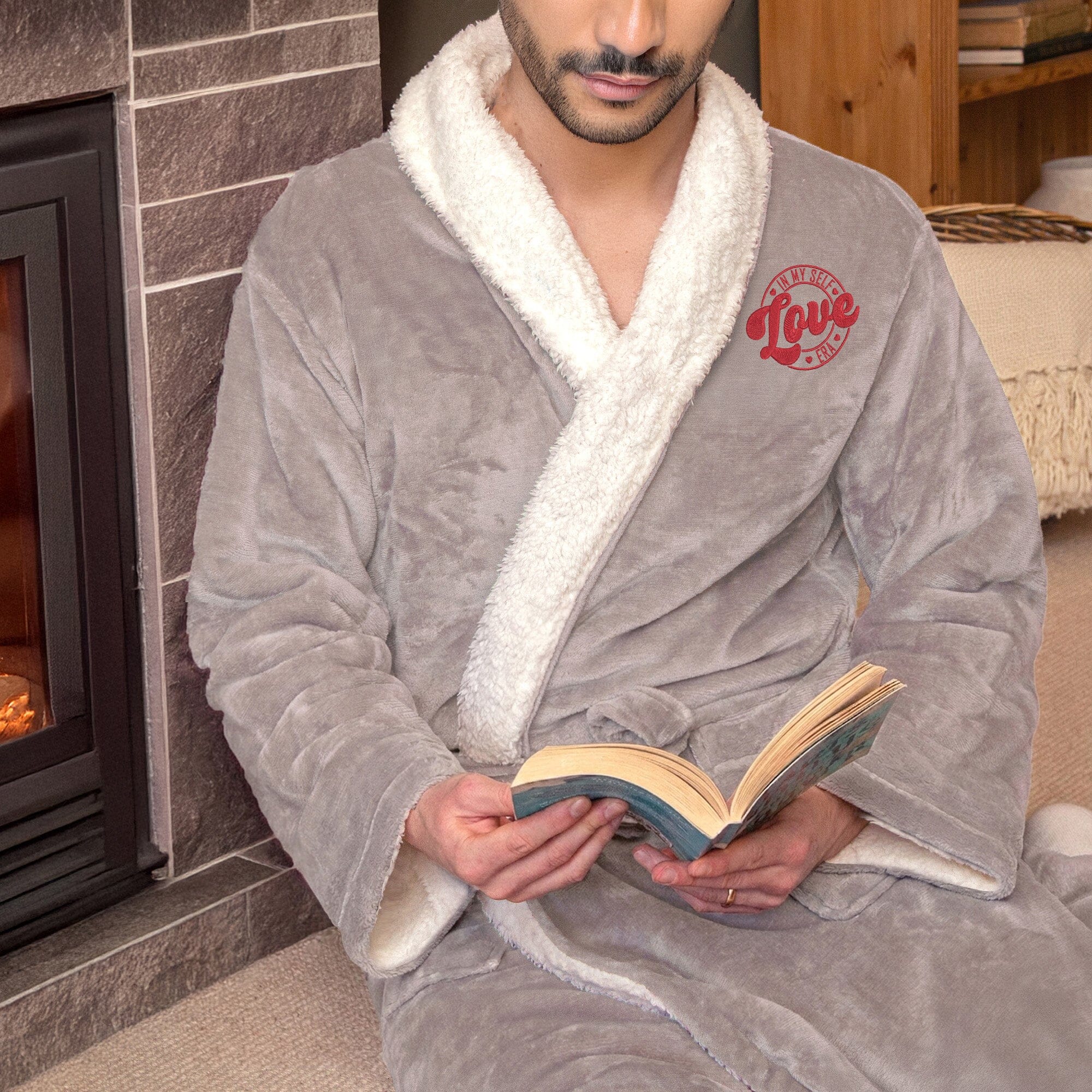 Man wearing an embroidered Taupe sherpa robe with 'Self Love Era' logo, sitting next to a fireplace