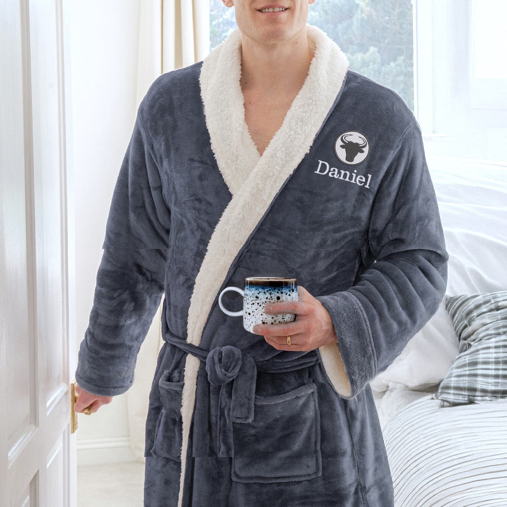 Person wearing a personalised zodiac slate robe with white trim, holding a mug, in a bedroom setting.