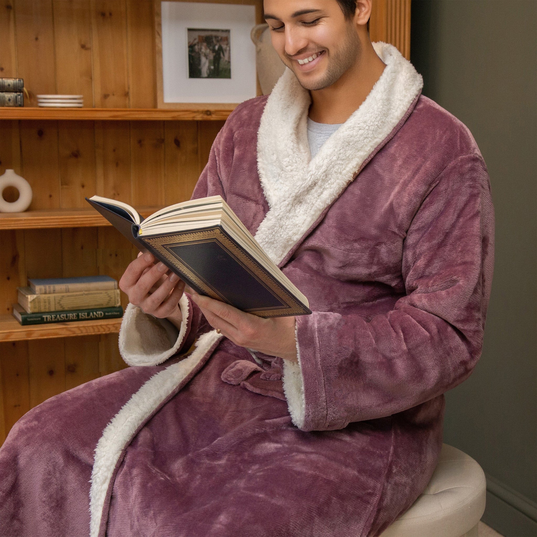 Man in a lavender sherpa robe with white trim reading a book in a cosy room.