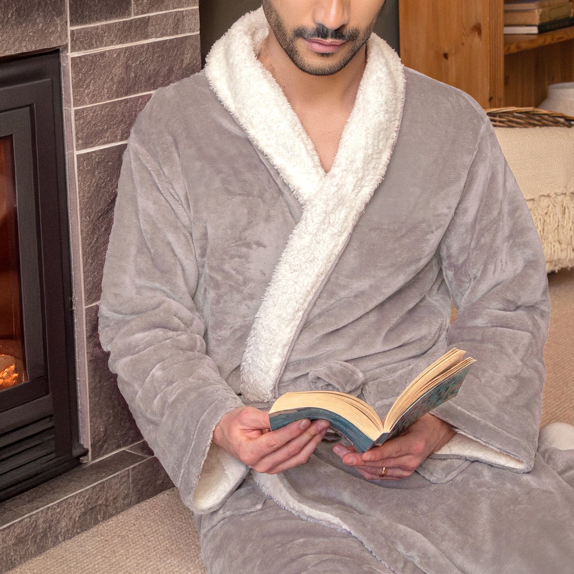 Man in a sherpa Taupe robe with white trim, reading a book by a fireplace.