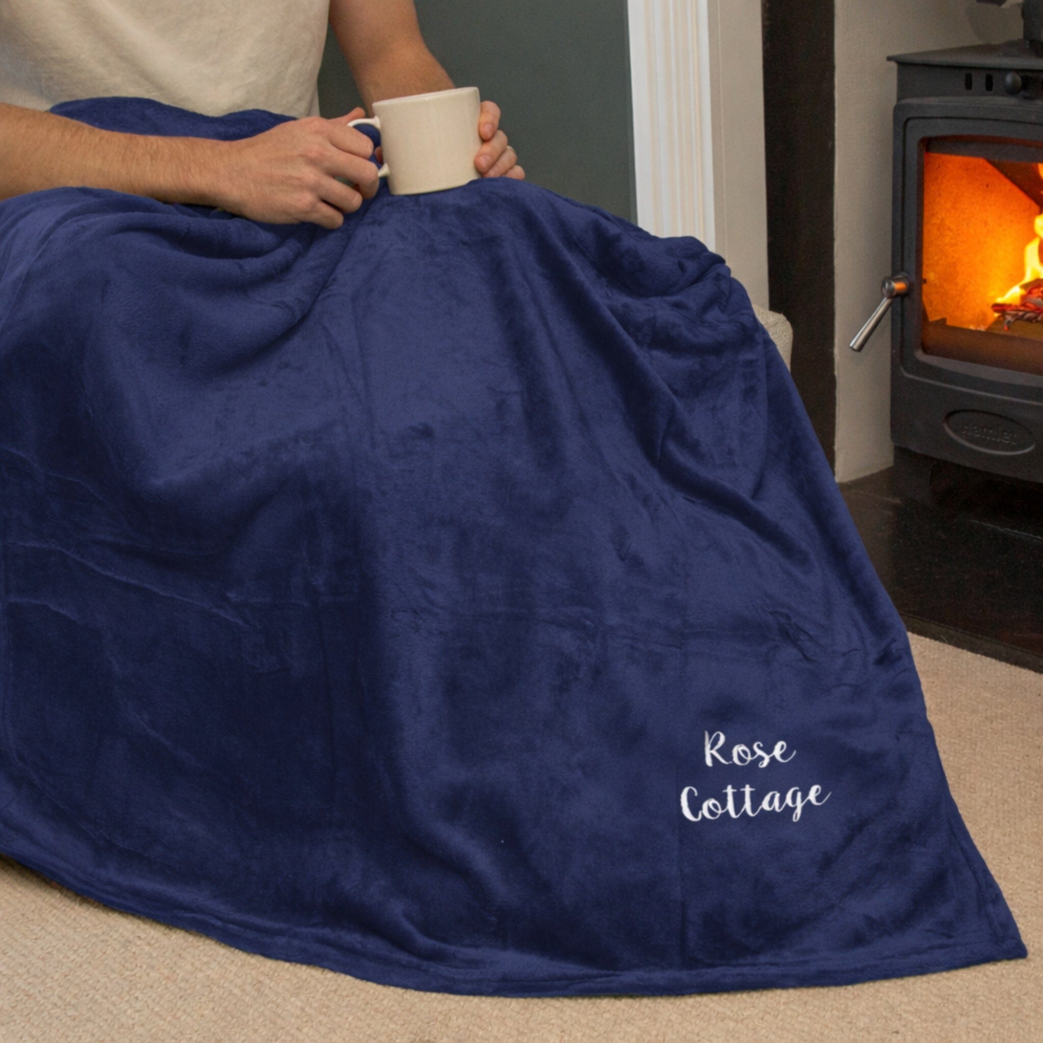 Person wrapped in a blue blanket with 'Rose Cottage' text, holding a mug by a fireplace.