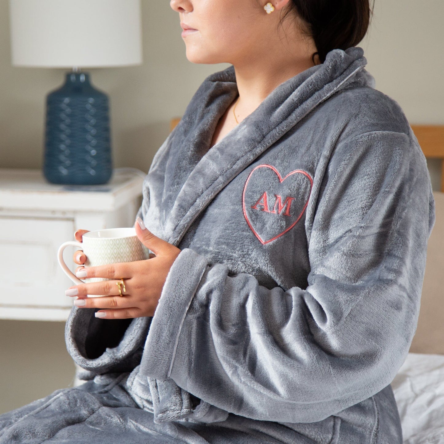 A female model sitting on a bed holding a mug wearing a Personalised Super Soft Star Monogrammed Robe in slate