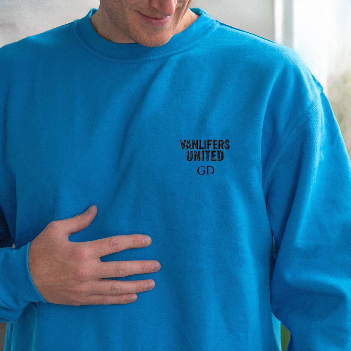 A close-up of a person wearing a bright blue crewneck sweatshirt with black text on the left chest reading 'VANLIFERS UNITED' and 'GD'.