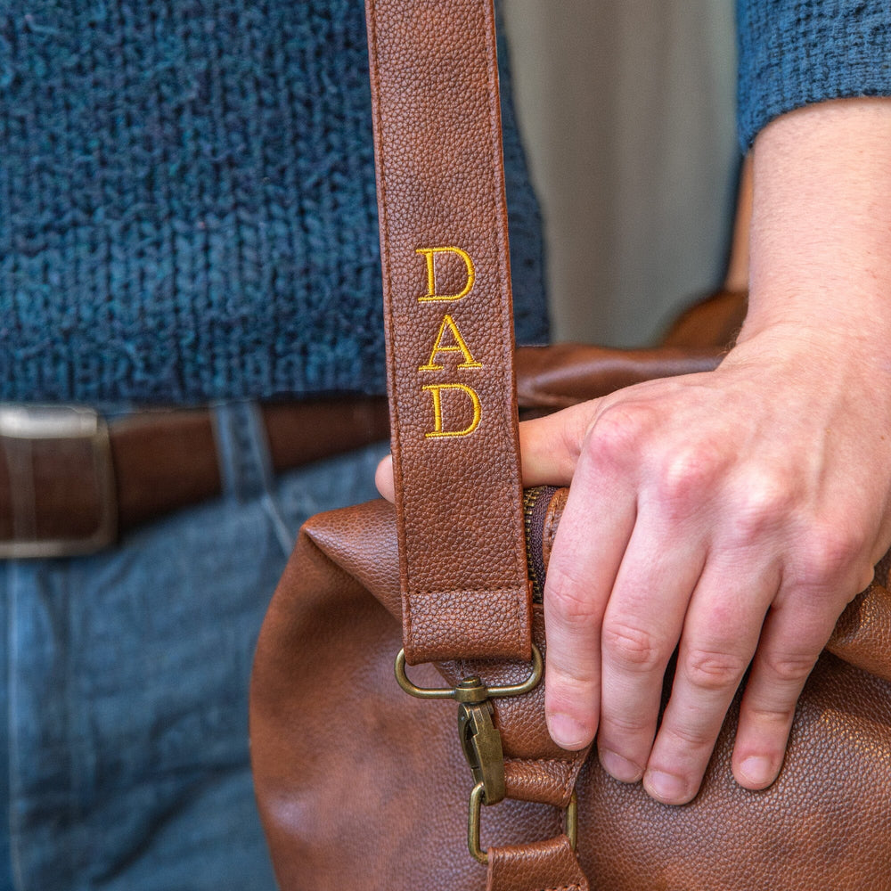 Brown leather bag with 'DAD' strap held by a person wearing a blue shirt.