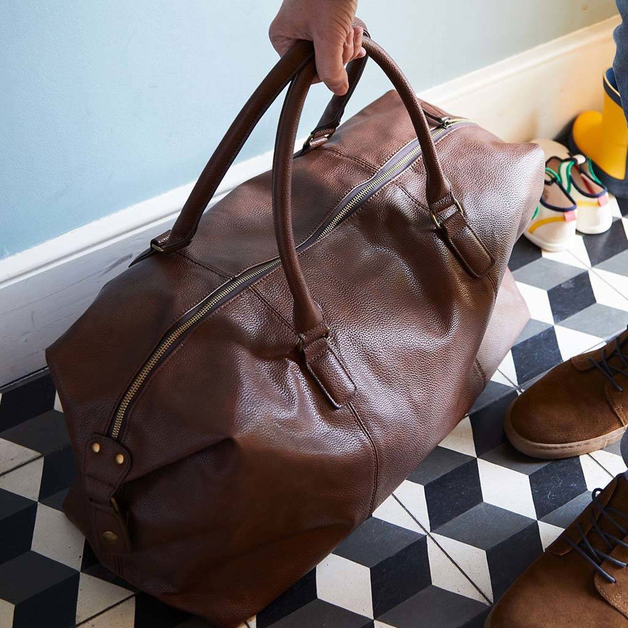 Brown leather duffel bag being held on a checkered floor with shoes nearby