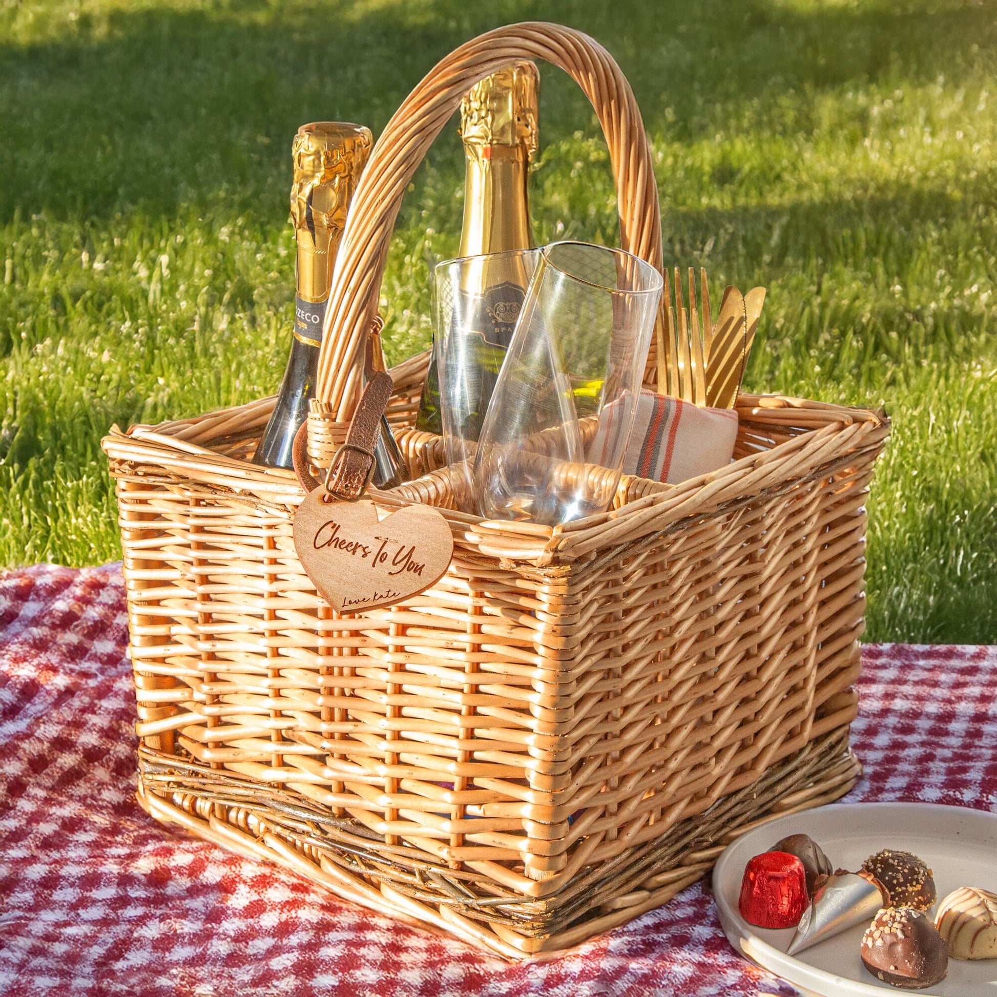 A personalised wicker wine carrier basket is in a picnic scene. Featuring a heart-shaped personalised cherry wood veneer tag