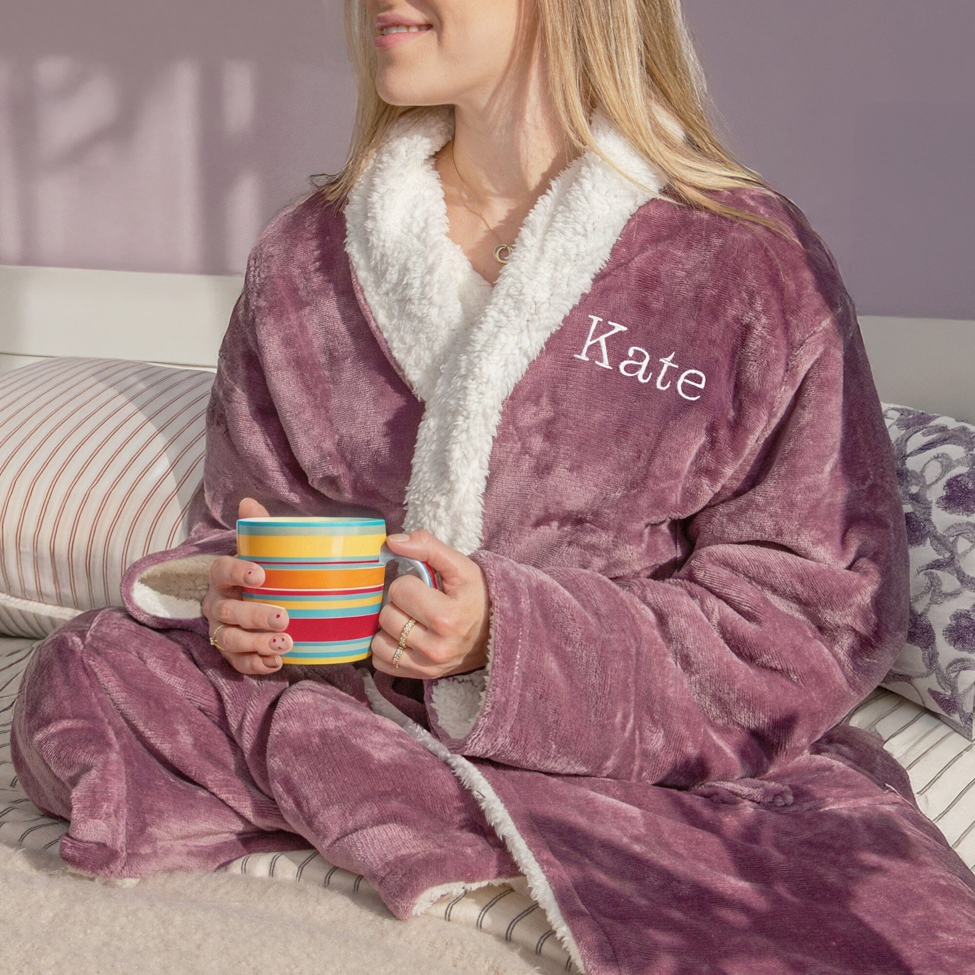 A female wearing a personalised lavender robe with 'Kate' embroidered on it, holding a colourful mug.