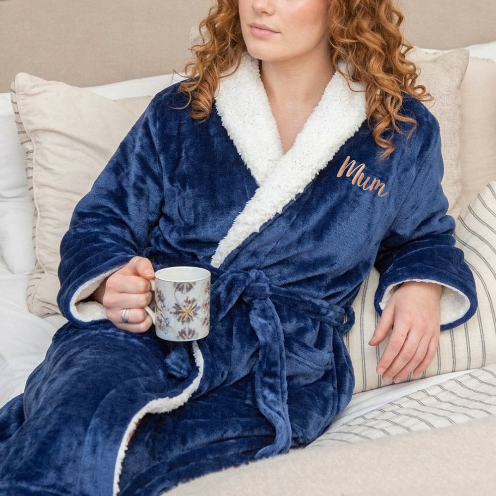 Woman in a blue robe with 'Mum' embroidered on it, holding a mug on a couch.